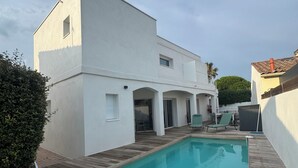 En modern vit byggnad med en swimmingpool, ett trädäck och utemöbler.