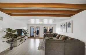 A living room with a sofa, a TV, and a plant. There are two doors leading to a balcony. - Spacious 5BR Home w/ Pool + Tiki Lounge (Hollywood)