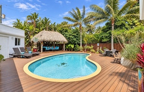 Ein Swimmingpool mit Holzterrasse, Liegestühlen, einer strohgedeckten Struktur und tropischer Vegetation.