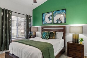 Un dormitorio con una cama, una mesita de noche, una lámpara, un ventanal con cortinas y dos cuadros colgados en la pared.