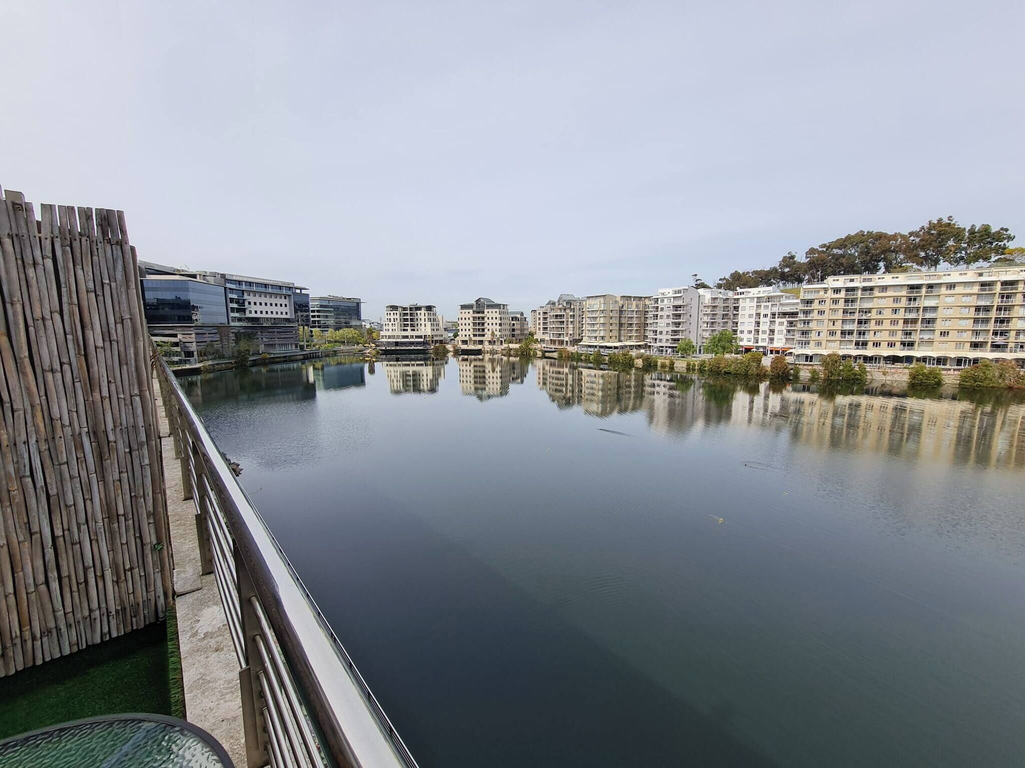 A calm water body reflecting buildings, a bamboo fence, and modern architecture.
