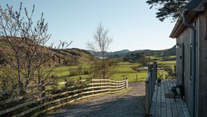 Un sendero de grava que conduce a una edificación de madera con un porche, rodeada por una cerca de madera y un paisaje rural con colinas y árboles.