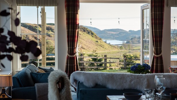 Una habitación con vistas a colinas y un lago, un sofá con una manta de piel, una mesa de comedor con vasos y un cuenco, y una lámpara sobre una mesita auxiliar.