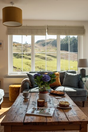 Ein gemütliches Wohnzimmer mit einem Holztisch, einer Couch und Blick auf sanfte Hügel durch große Fenster.
