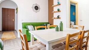 Zona pranzo con tavolo bianco, sedie di legno, divano verde e orologio da parete geometrico.