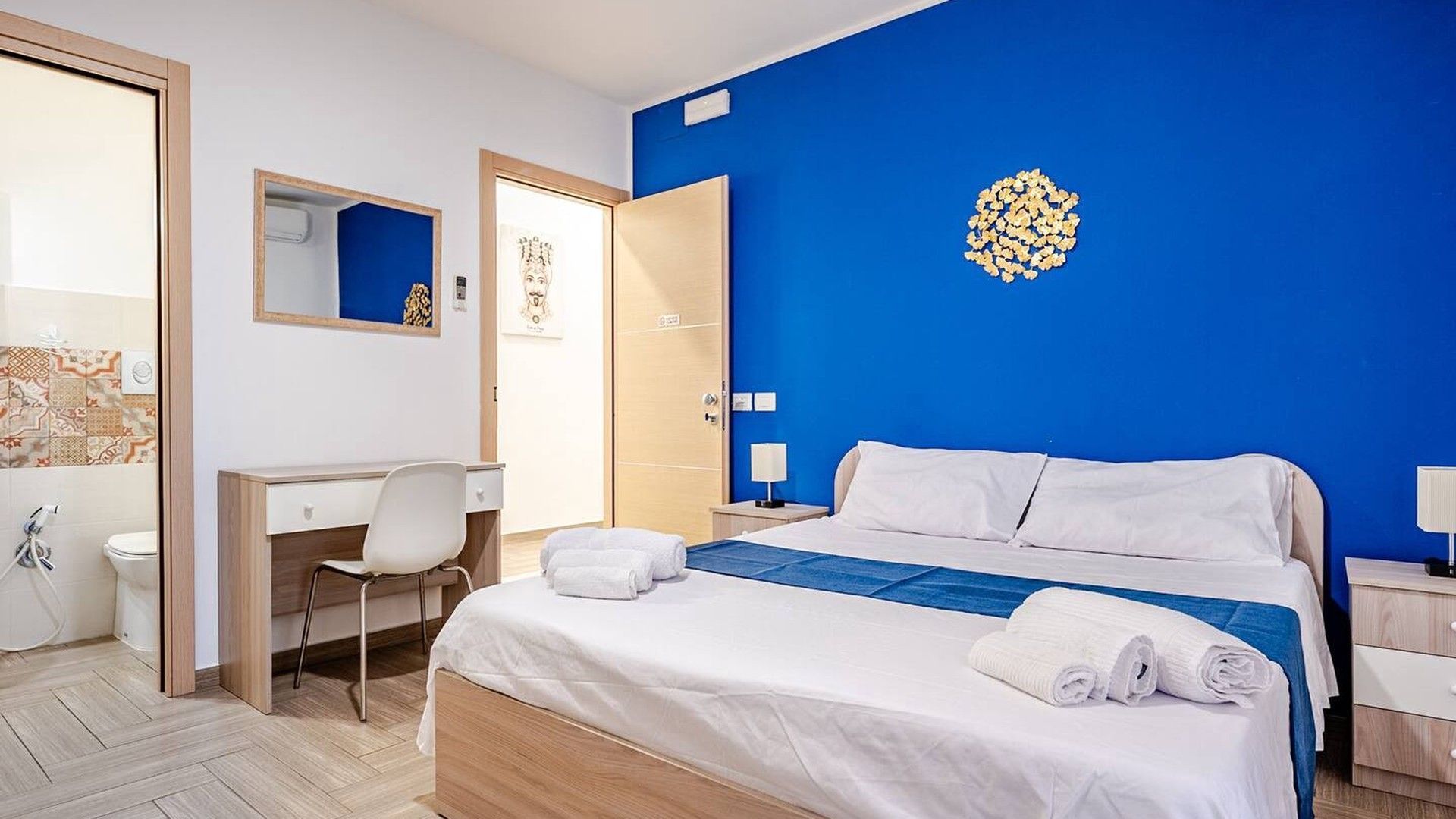 A hotel room with a bed, a desk, a chair, a bathroom, and a blue accent wall.