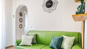 Un divano verde con cuscini, una sveglia da parete con decorazioni geometriche e una mensola in legno con un vaso verde.