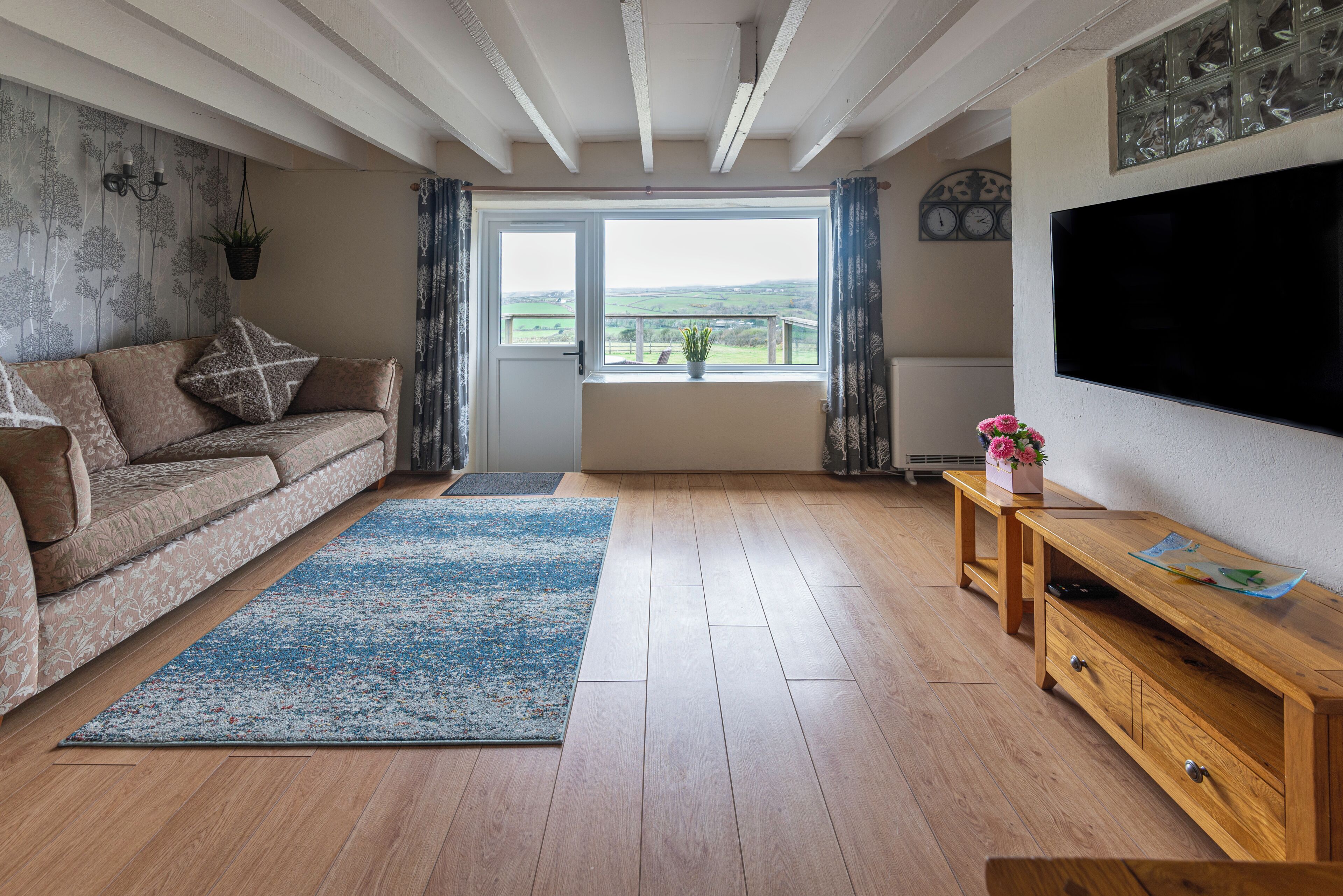 A living room with a sofa, a wooden coffee table, a TV, a large window with curtains, and a view of the countryside.