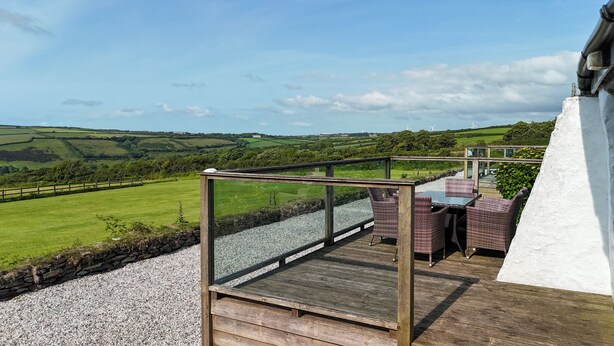 A terrace with a table and chairs, overlooking a green landscape with wind turbines in the distance. - Courtyard Farm Cottages (Boscastle)