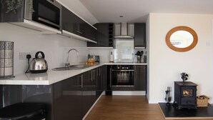A modern kitchen with black cabinets, a microwave, a kettle, and a wood-burning stove. - Courtyard Farm Cottages (Boscastle)