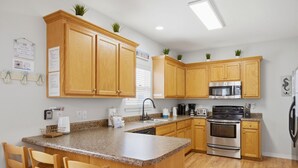 A kitchen with wooden cabinets, granite countertops, stainless steel appliances, and a central island with bar stools. - Rookery III 6005 - Peach and Quiet (Gulf Shores)