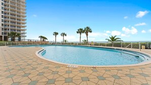 A swimming pool with a tiled deck, surrounded by palm trees and a high-rise building. - Beach Colony East 7B (Pensacola)