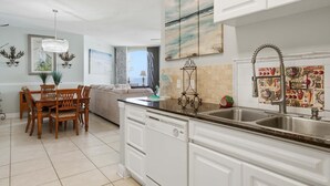 A modern kitchen with white cabinetry, a double sink, and a dining area with a wooden table and chairs. - Beach Colony East 7B (Pensacola)