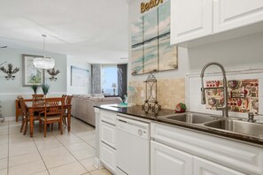 A modern kitchen with white cabinetry, a double sink, and a dining area with a wooden table and chairs. - Beach Colony East 7B (Pensacola)