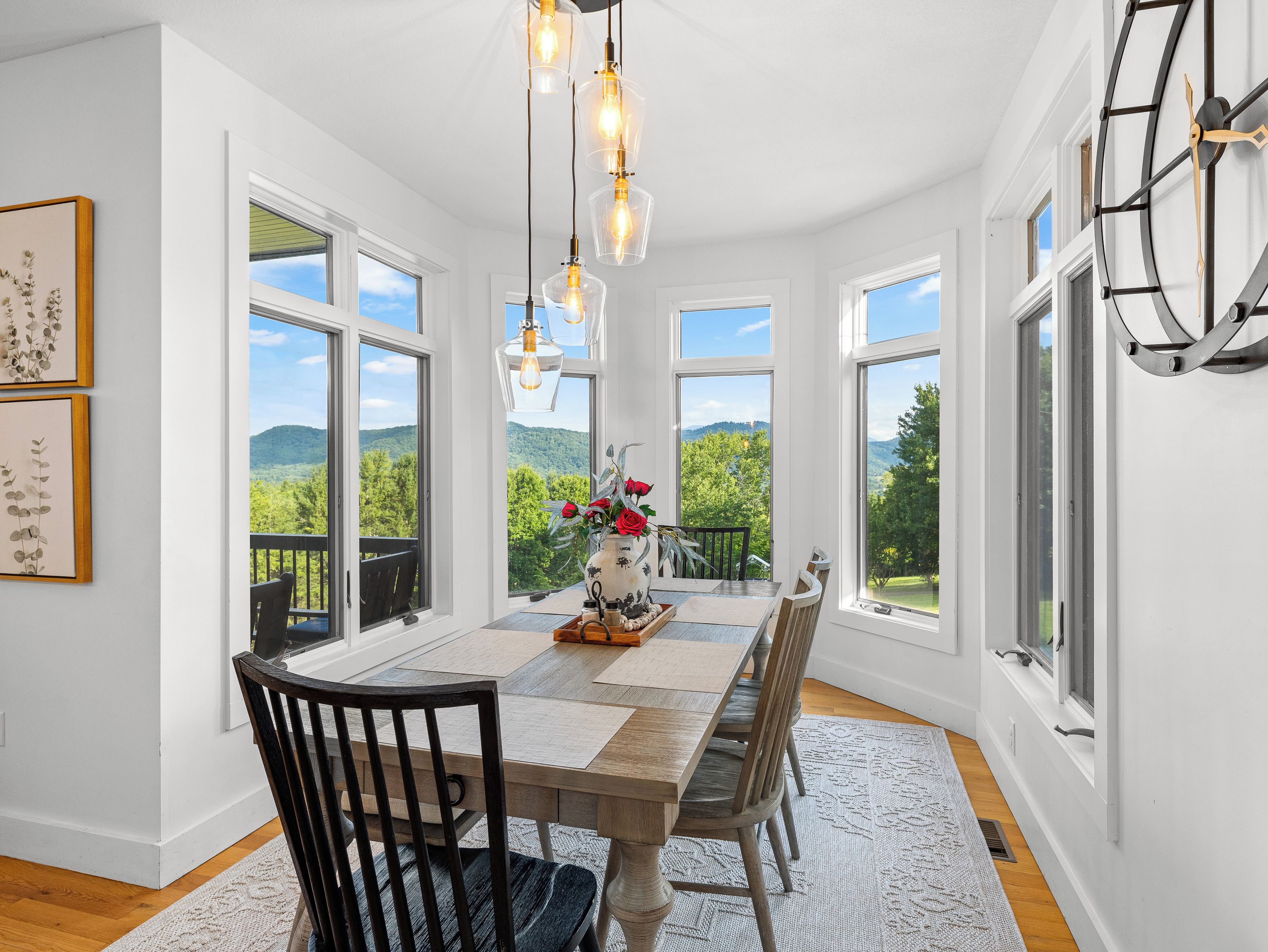 Ein Essbereich mit Holztisch, Stühlen und einer Blumenvase. Große Fenster bieten Blick auf Grünflächen und Hügel.