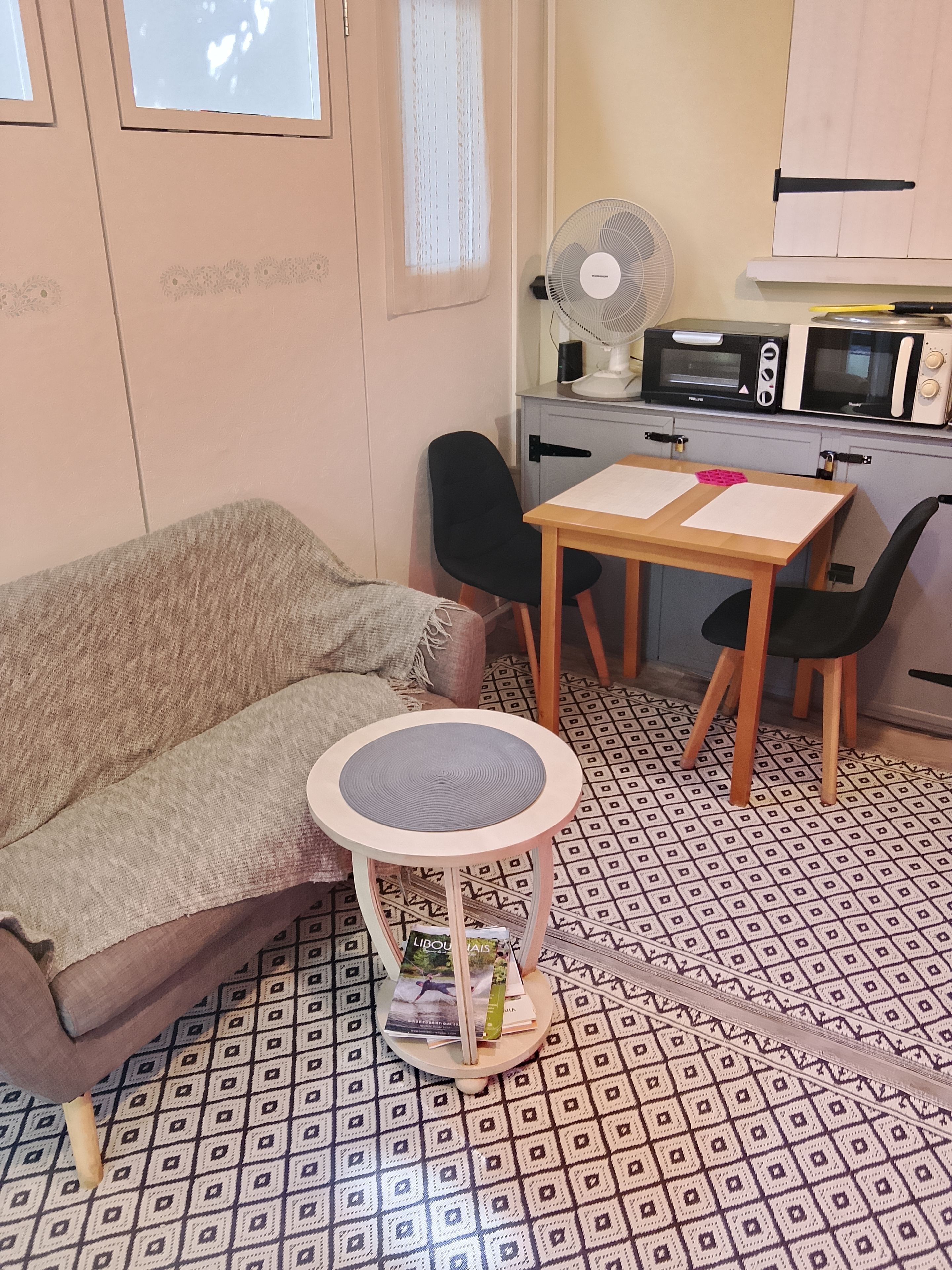 Une petite cuisine équipée d’un four à micro-ondes, d’un grille-pain et d’un ventilateur. On y trouve une table avec des chaises et une petite table ronde sur laquelle sont posés un livre et un magazine. La pièce est dotée d’un revêtement de sol à motifs et d’un canapé.