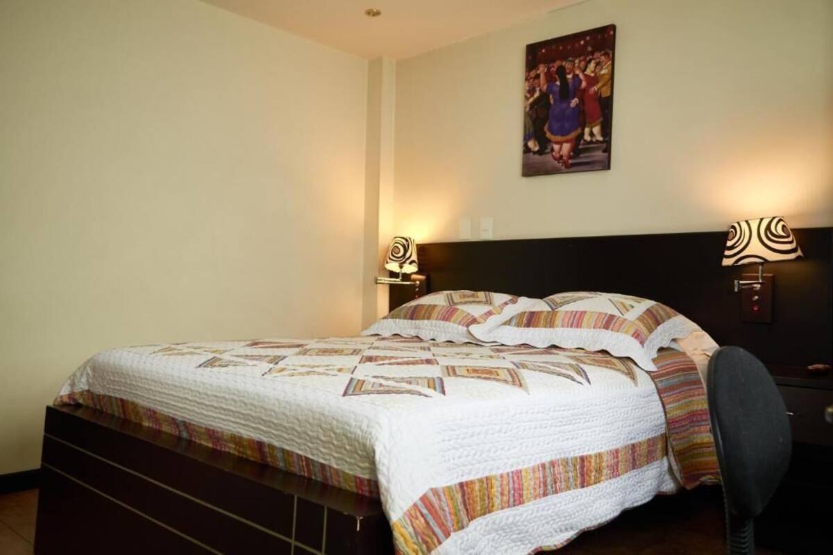 Una habitación de hotel con cama, lámparas de noche, mesita de noche y un cuadro colgado en la pared.