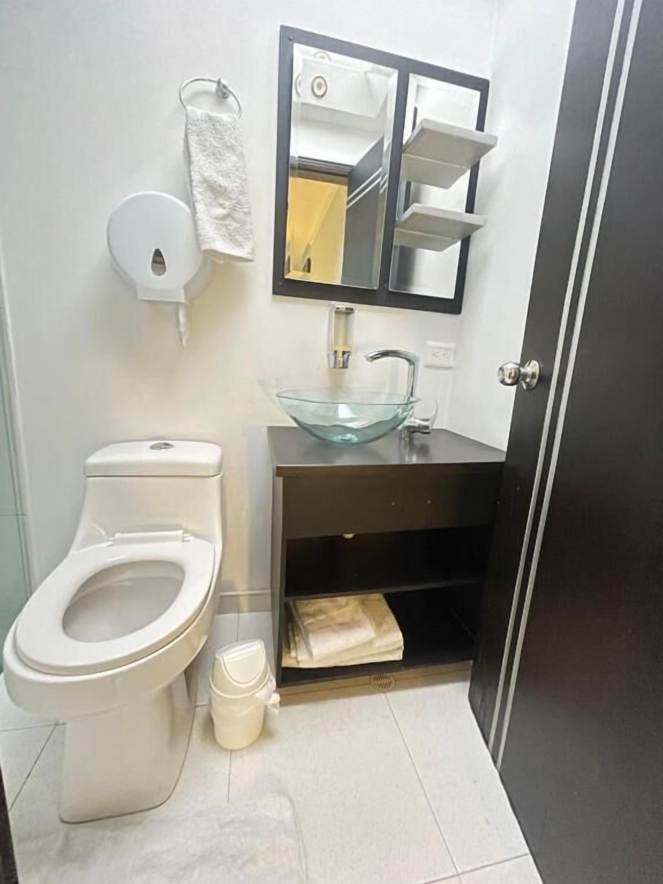 Une salle de bain équipée de toilettes, d’un lavabo en verre, d’un miroir et d’un distributeur de serviettes.