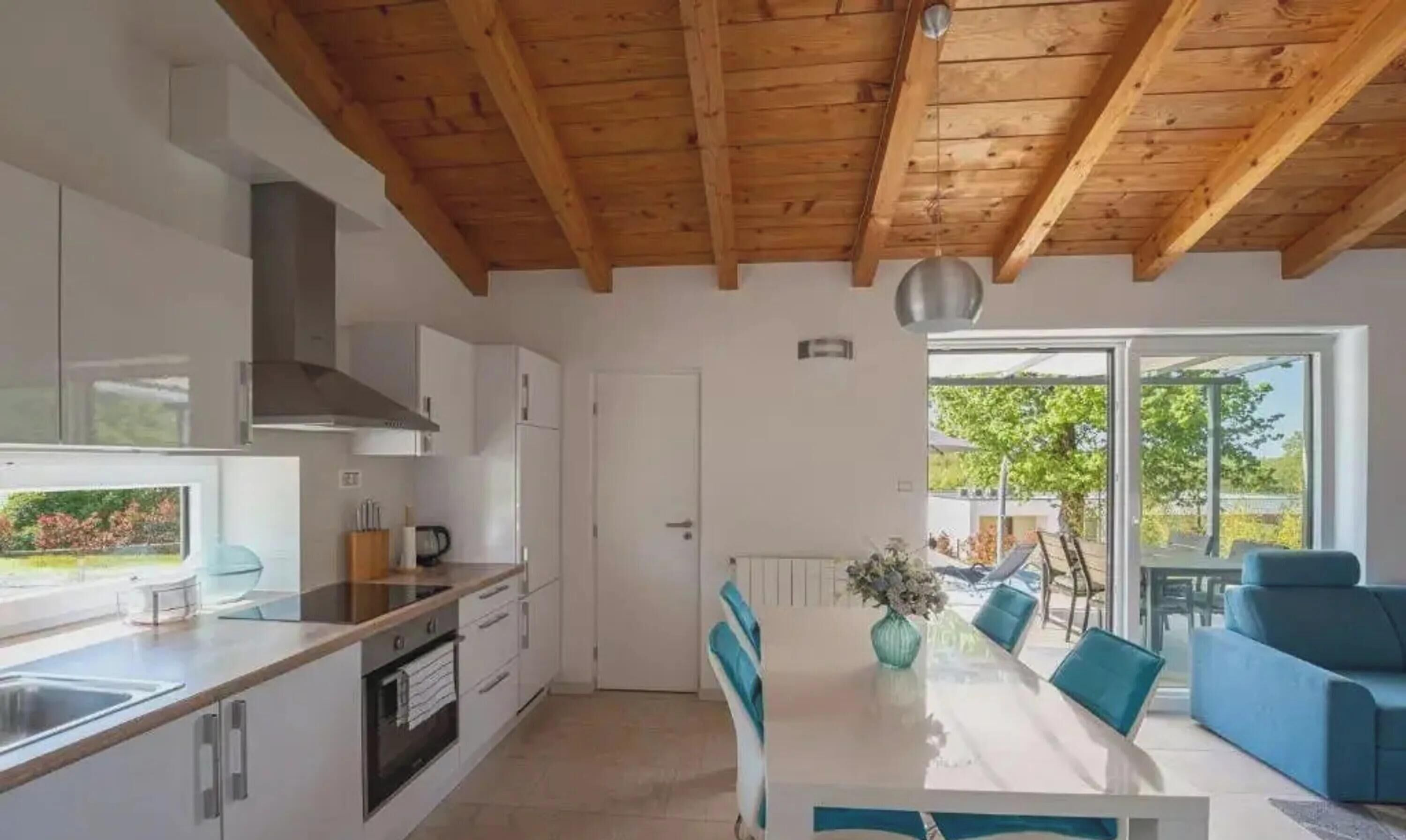 Cozinha moderna com vigas de madeira, área de jantar com mesa e cadeiras, e vista através de uma porta de vidro.