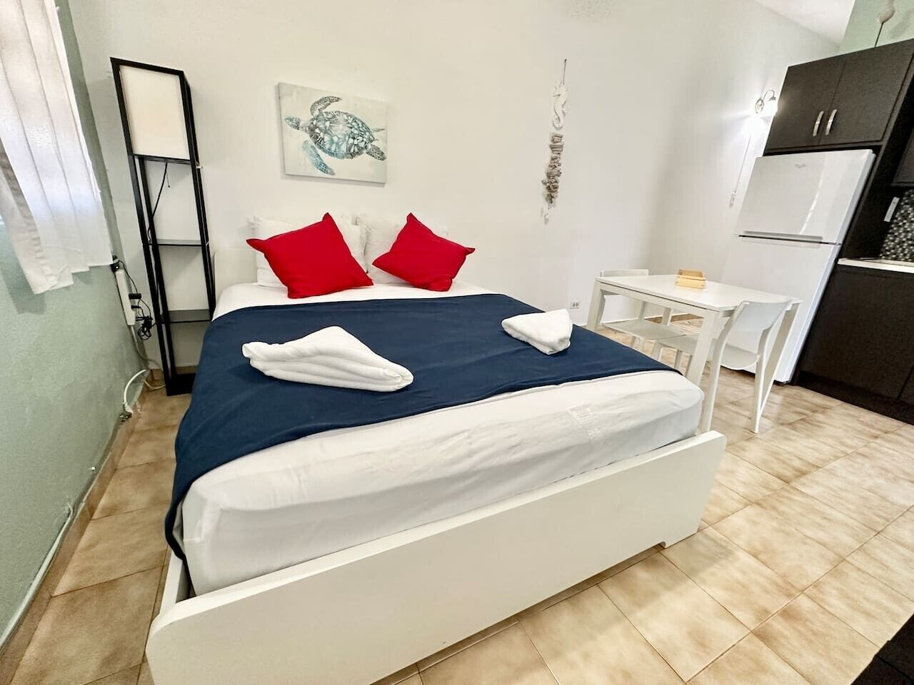 Ein Schlafzimmer mit einem Bett, einem Nachttisch, einem kleinen Schreibtisch, einem Kühlschrank und einem an der Wand befestigten Kunstwerk.