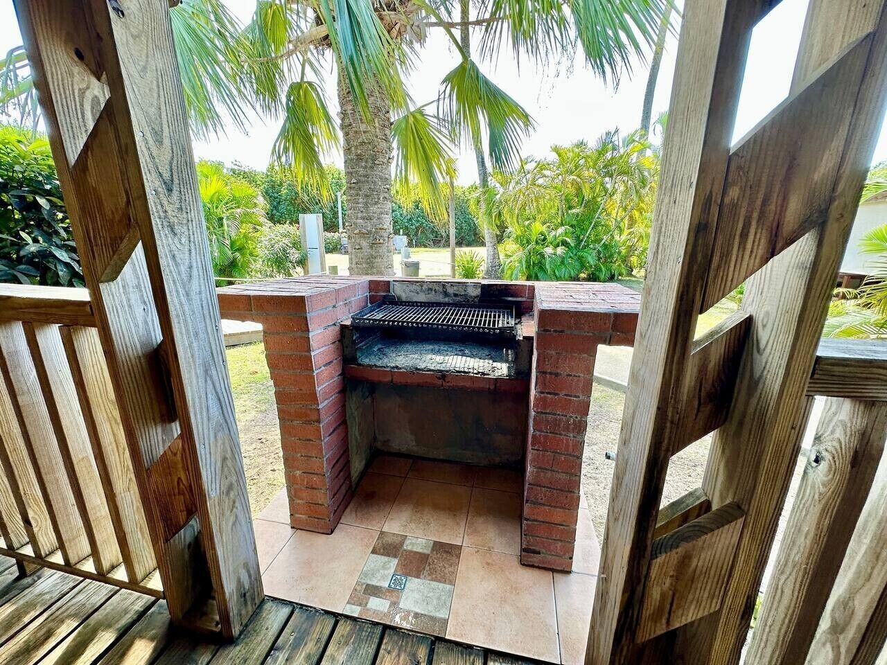 A barbecue grill with a brick base, surrounded by a wooden fence and palm trees.