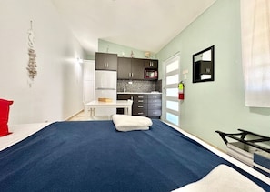A bedroom with a large bed, a desk, a chair, a wardrobe, and a kitchenette.