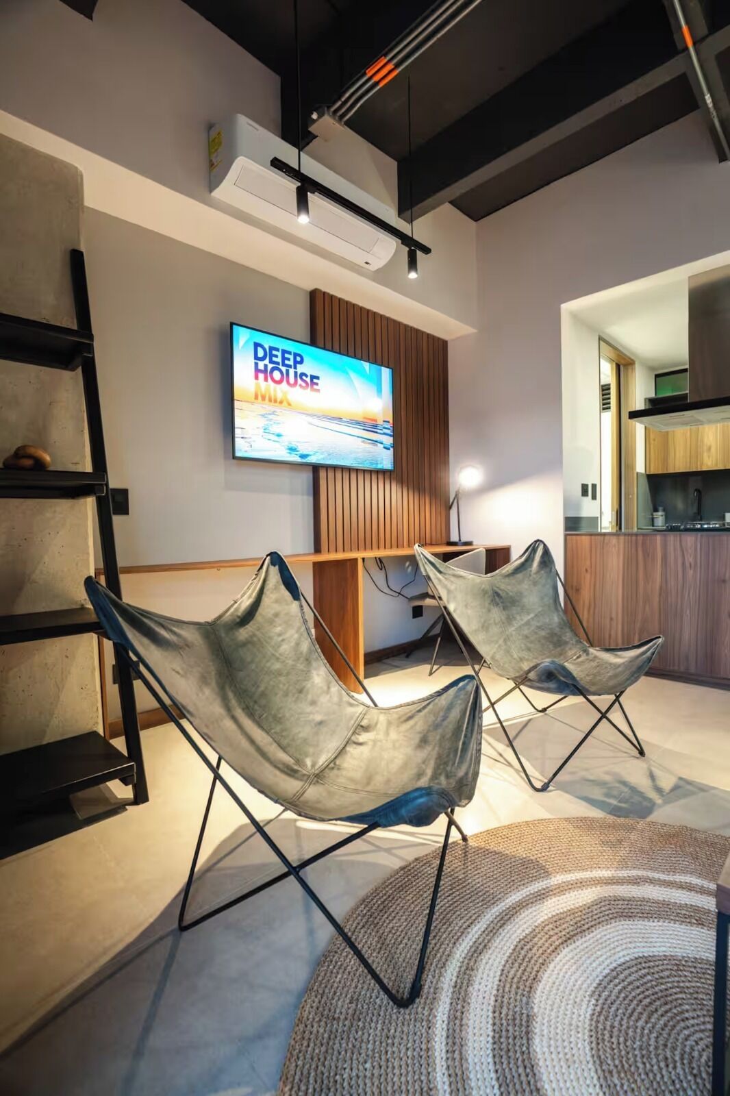 A modern living room with a flat-screen TV, two leather chairs, a wooden accent wall, and a kitchen area in the background.
