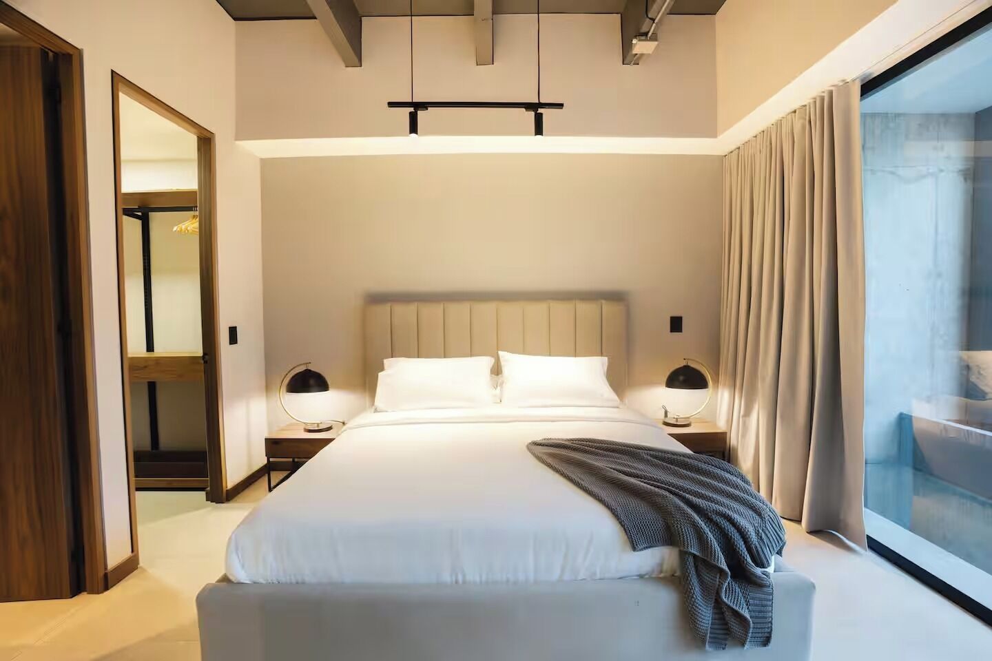 A modern hotel room with a large bed, bedside lamps, a view through a window, and a wooden door.
