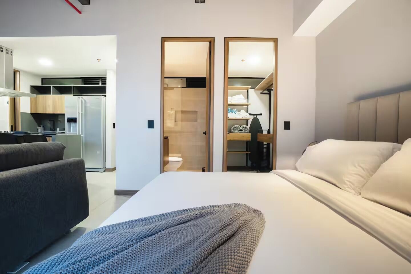 A modern hotel room with a large bed, a sofa, a kitchenette, and a bathroom visible through a glass door.