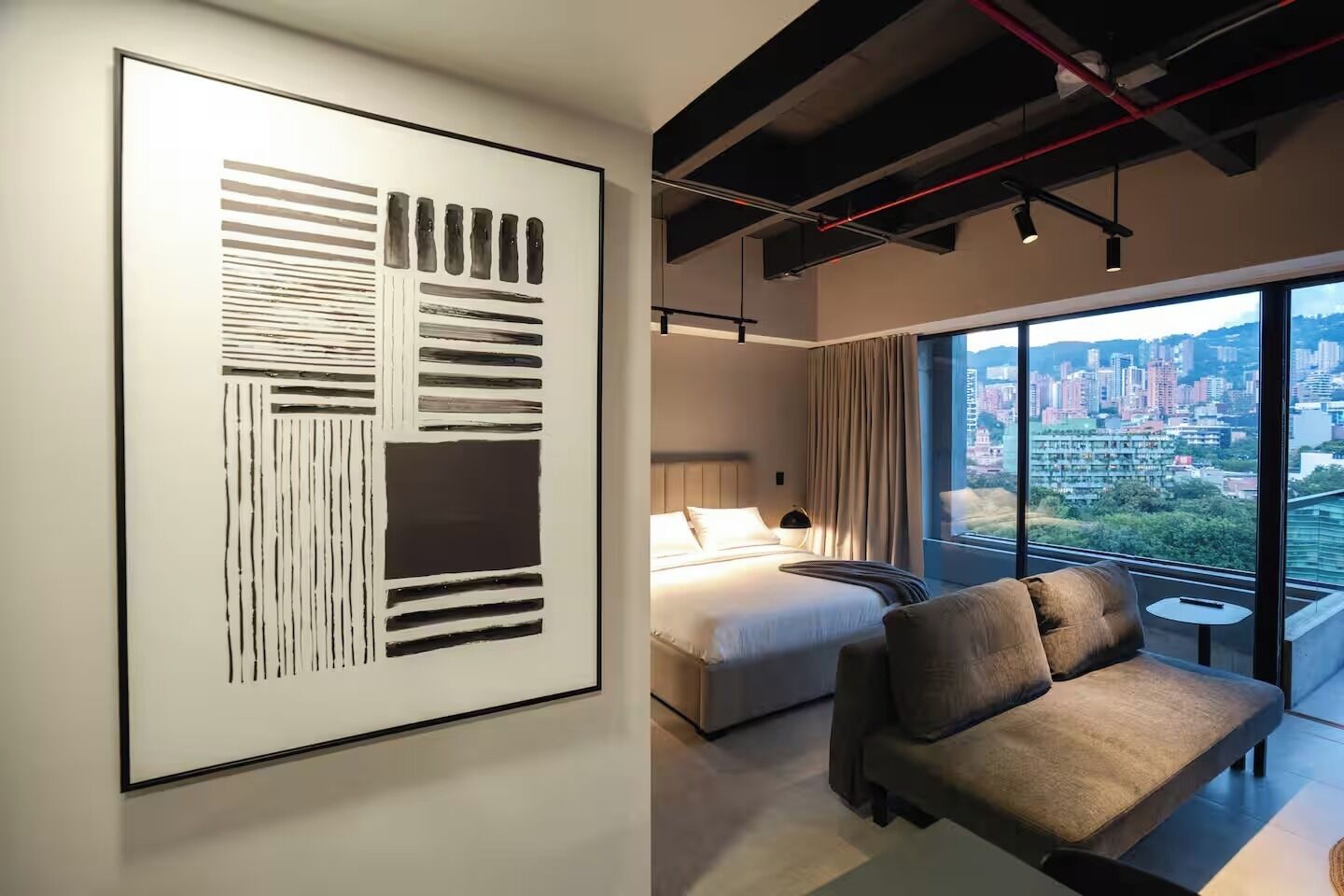 Habitació d'hotel moderna amb un quadre abstracte gran, un llit, un sofà i vista a la ciutat a través de grans finestres.
