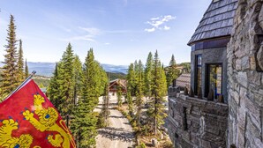 Ein Steinhaus mit Holzdach, eine Flagge mit Löwenmustern und Blick auf eine Hütte in einem bewaldeten Gebiet.