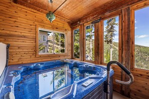 A wooden cabin with a hot tub, large windows, and a view of the forest.
