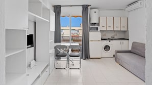 Un espacio habitable compacto con kitchenette, lavadora, un sofá gris y una zona de comedor con una silla.