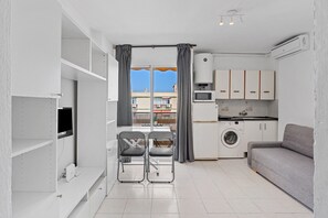 A compact living space with a kitchenette, a washing machine, a grey sofa, and a dining area with a chair. - 'Las Naciones' Studio with Sea Views, Wi-Fi, and Air Conditioning (Benalmádena)