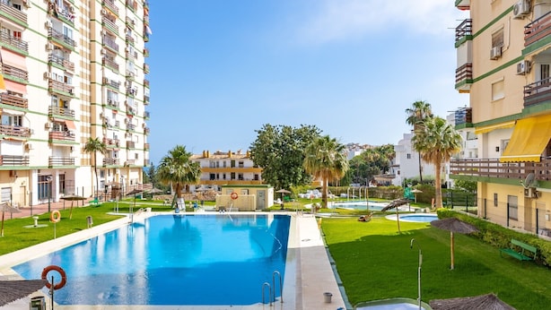 Una piscina rodeada de césped cuidado y palmeras, con edificios altos al fondo.