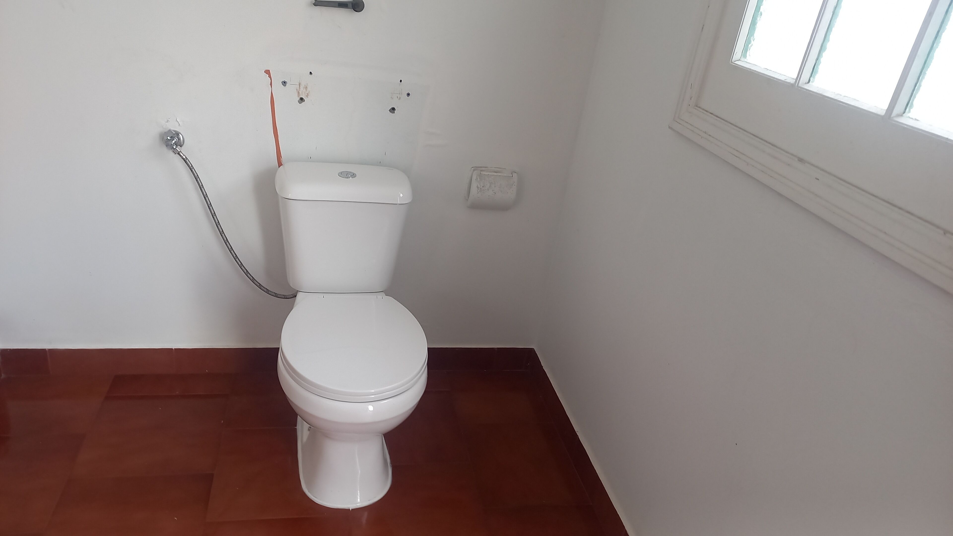 Un inodoro blanco con tanque, una tubería visible y una ventana con persianas.
