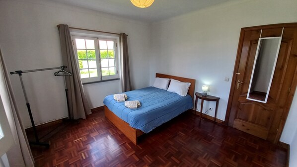 A bedroom with a wooden bed, a bedside table with a lamp, a window with curtains, and a wooden floor. - Alojamento Pascoal (Ribeira Grande)