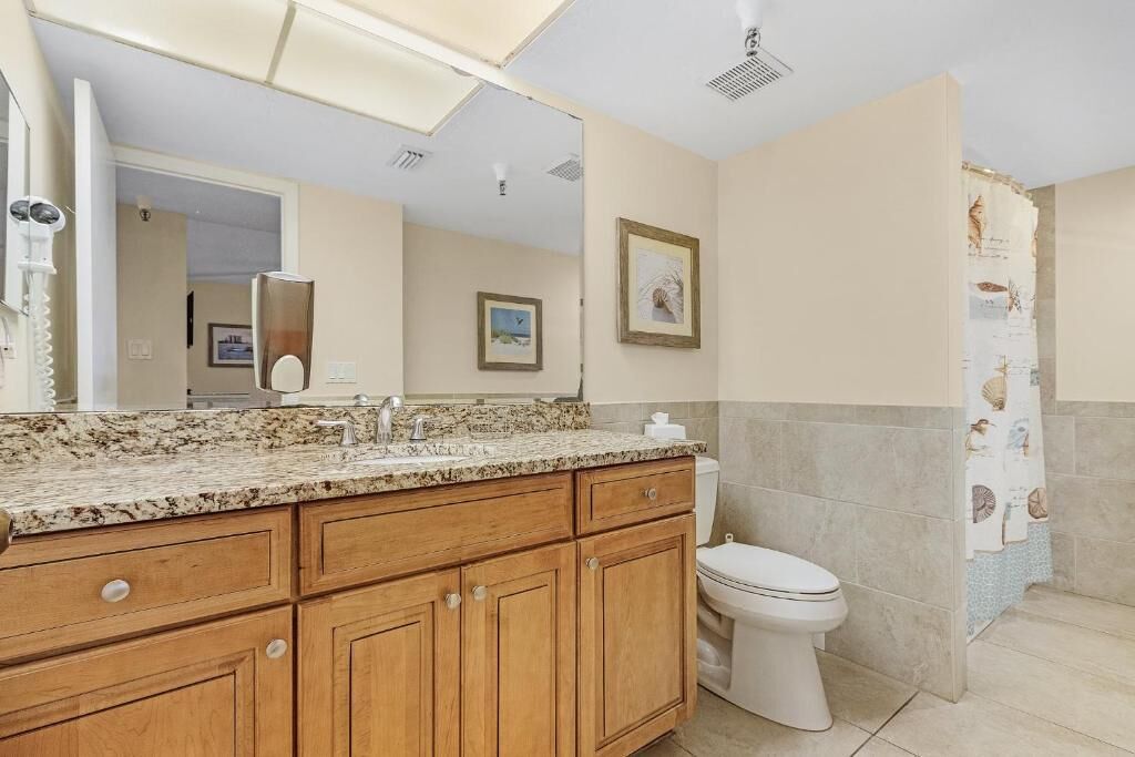 A bathroom with a granite countertop, wooden cabinets, a white toilet, and a shower curtain with a nautical theme.