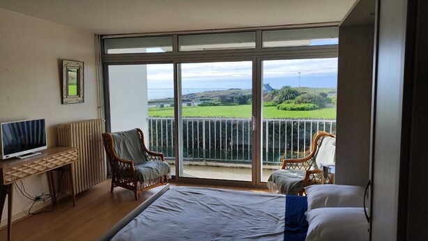 A bedroom with a large window, a bed, a TV, two chairs, and a radiator. - Grand studio Résidence les 3 Iles Quiberon N°28 (Quiberon)