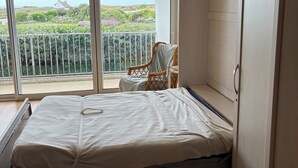 A room with a bed, a chair, and a view of a balcony with greenery and a distant building. - Grand studio Résidence les 3 Iles Quiberon N°28 (Quiberon)