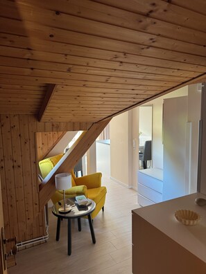 Een kamer met houten balken in het plafond, een gele fauteuil, een klein rond tafeltje met een lamp en een wit kastje.