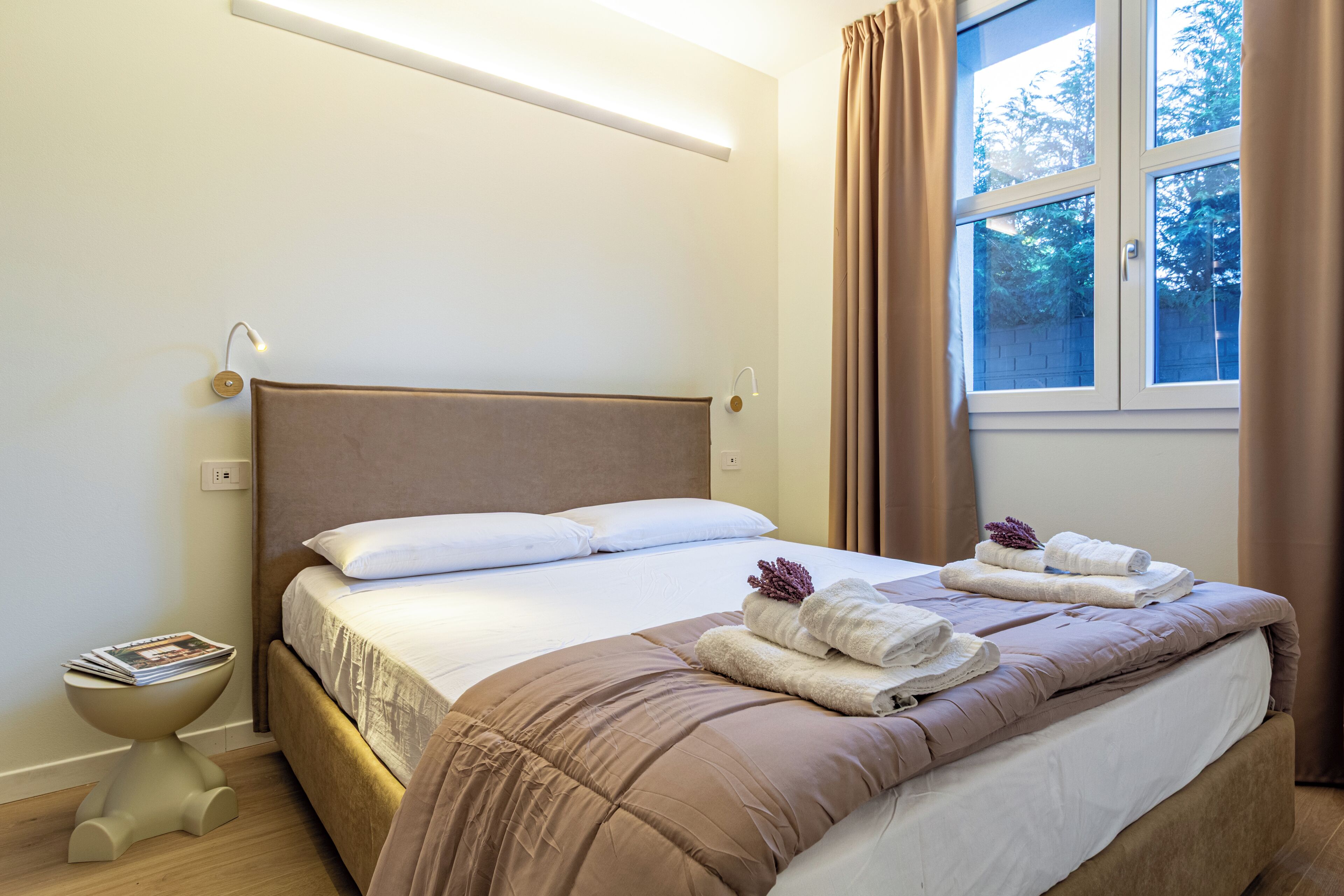 Una camera d'albergo con un letto, un comodino con una rivista e una finestra con tende.