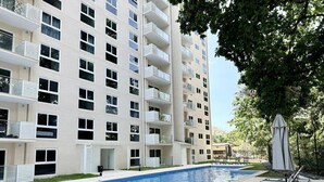 Une piscine avec des chaises longues, un immeuble à plusieurs étages doté de balcons et un ciel dégagé.