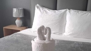 Un lit avec une couvre-lit grise, un porte-serviettes blanc orné d’un motif de cygne, une tête de lit grise et une lampe de chevet posée sur une table de nuit en bois.