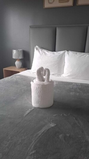 Ein Bett mit grauer Bettdecke, ein weißer Handtuchhalter mit Schwanenmotiv, ein grauer Kopfteil und eine Nachttischlampe auf einem Holznachttisch.