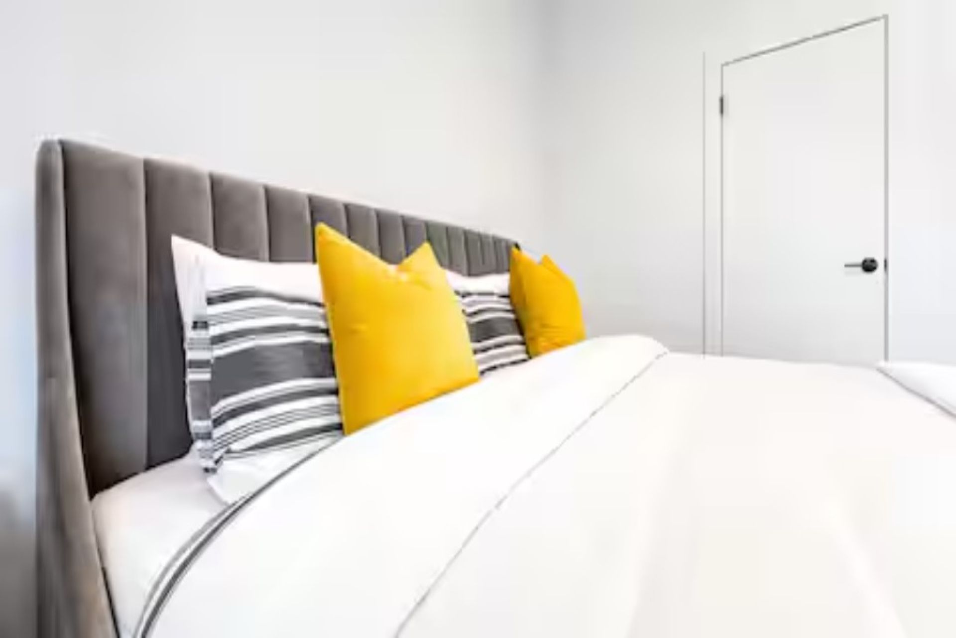 Un lit avec une tête de lit rembourrée de couleur grise, une literie blanche et deux oreillers jaunes.