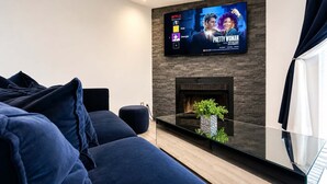 A modern living room with a flat-screen TV mounted on a brick wall, a glass coffee table, and a dark blue sofa. - Lakefront |Long Term Friendly |Pets|Cozy Fireplace (Jacksonville)