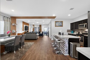 A modern kitchen with a central island, bar stools, and a dining area with a sofa and chairs. - Spacious Retreat | Pool Table, Firepit + Backyard (Houston)