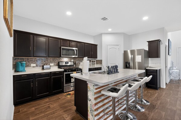 A modern kitchen with dark cabinetry, stainless steel appliances, a central island with bar stools, and a tiled backsplash. - Spacious Retreat | Pool Table, Firepit + Backyard (Houston)