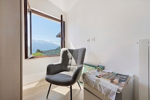 Una stanza con vista su un paesaggio montuoso attraverso una finestra, una poltrona grigia, un tavolino con una rivista e un muro bianco.